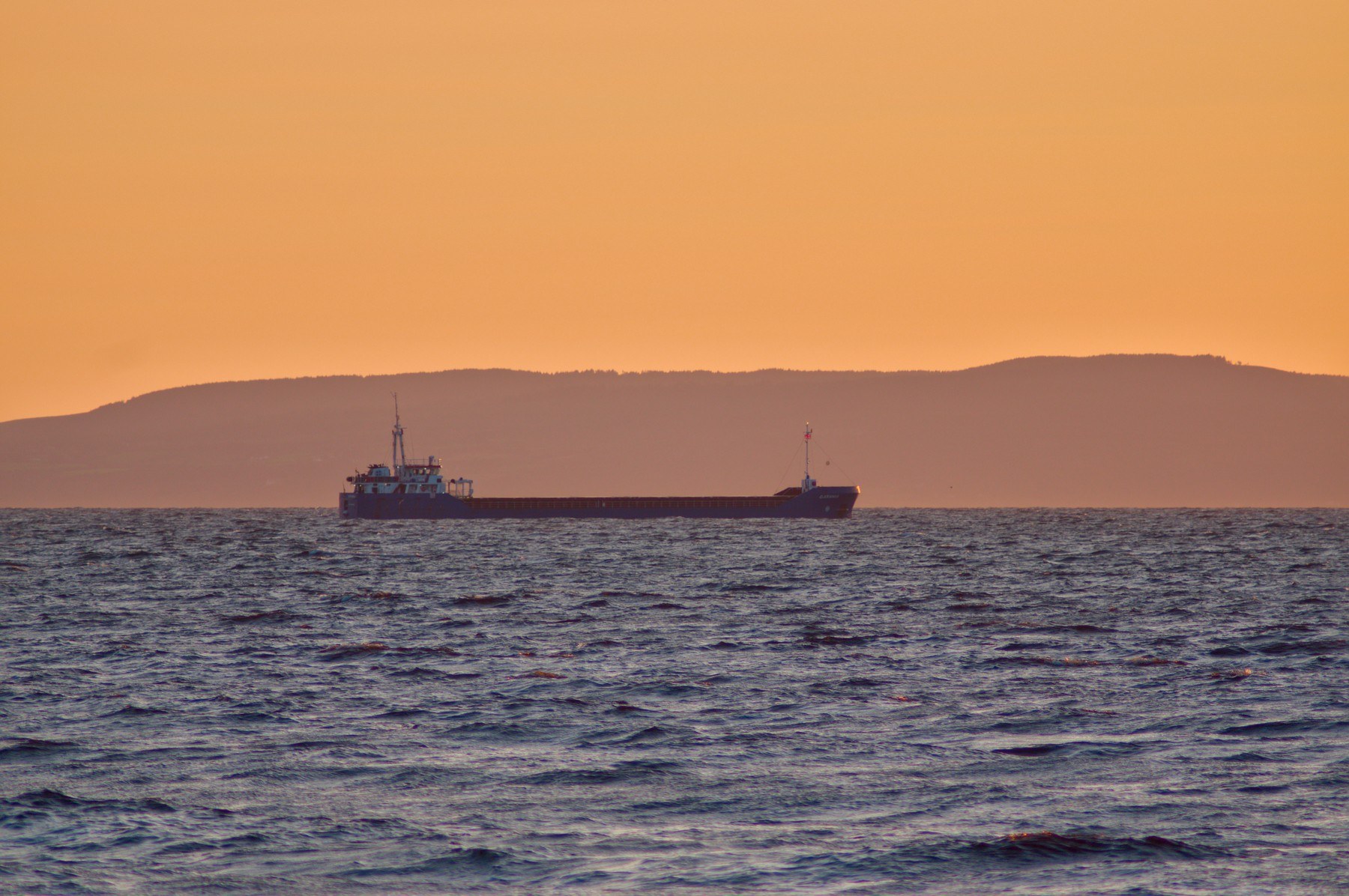 A cargo vessel on choppy sea, silhouetted before a low dark ridge beneath an orange sky.