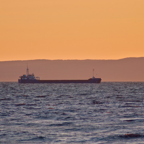 A cargo vessel on choppy sea, silhouetted before a low dark ridge beneath an orange sky.