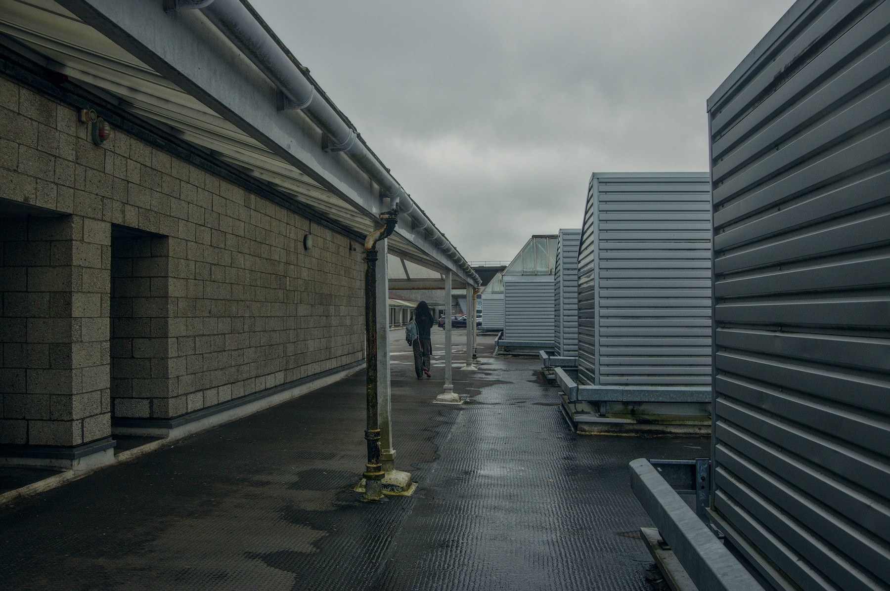 A lone pedestrian in a wet covered passage between a blockwork wall with drainpipes and tall corrugated metal screens at Cumbernauld Center.