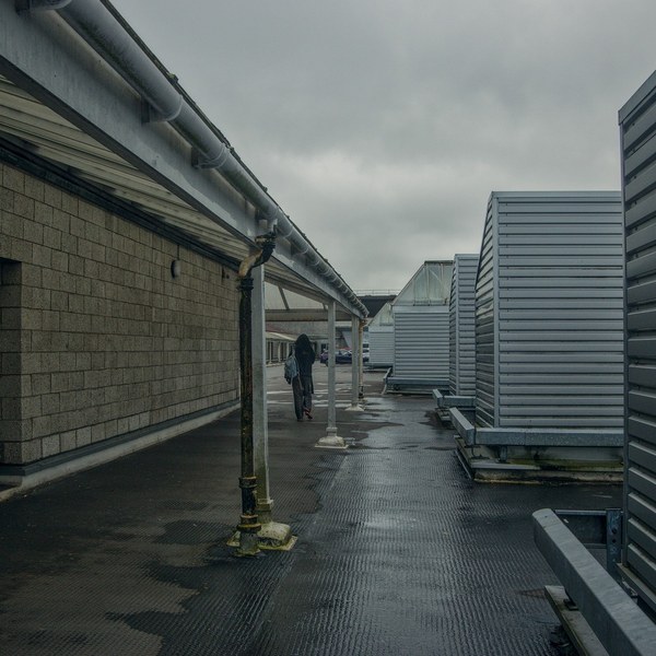 A lone pedestrian in a wet covered passage between a blockwork wall with drainpipes and tall corrugated metal screens at Cumbernauld Center.