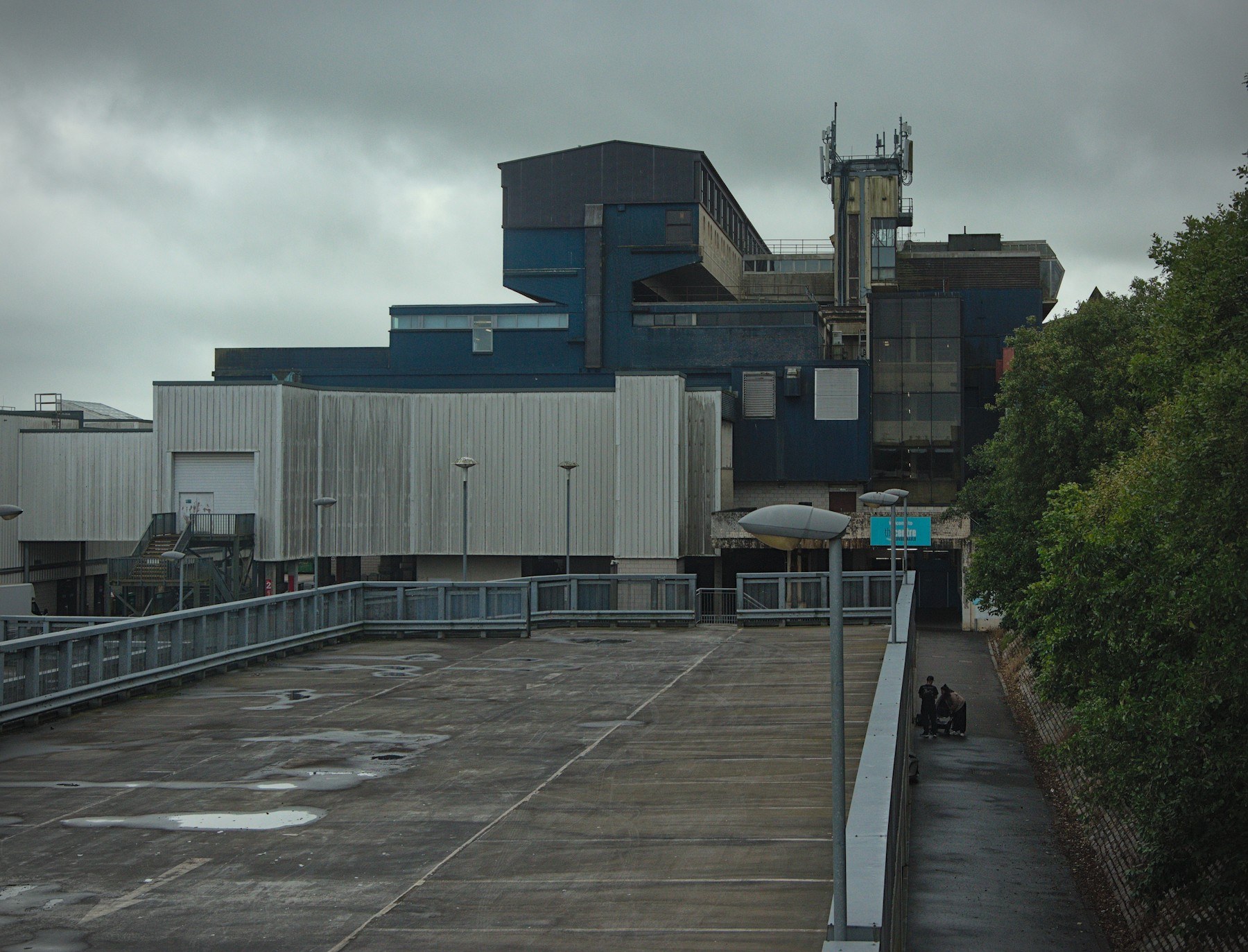 Rain-slicked paved forecourt leading to the layered blue and grey facade of Cumbernauld Center, with a communications mast above and two people on a covered path at right.