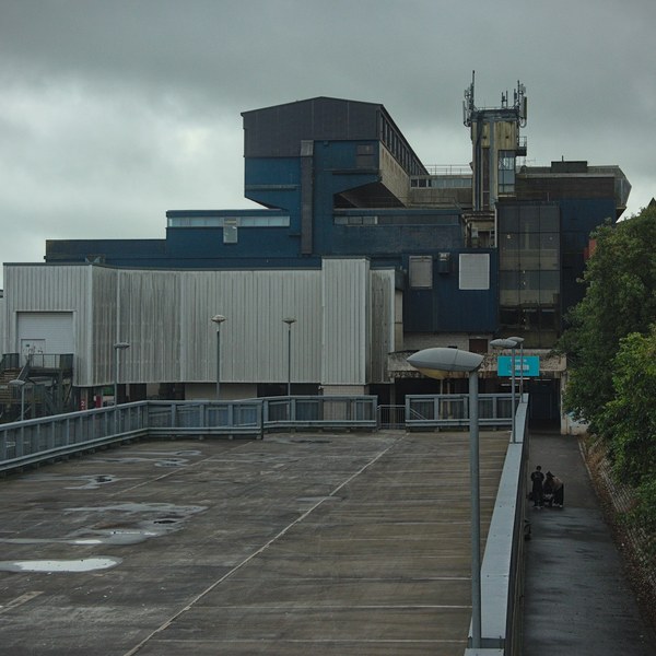 Rain-slicked paved forecourt leading to the layered blue and grey facade of Cumbernauld Center, with a communications mast above and two people on a covered path at right.