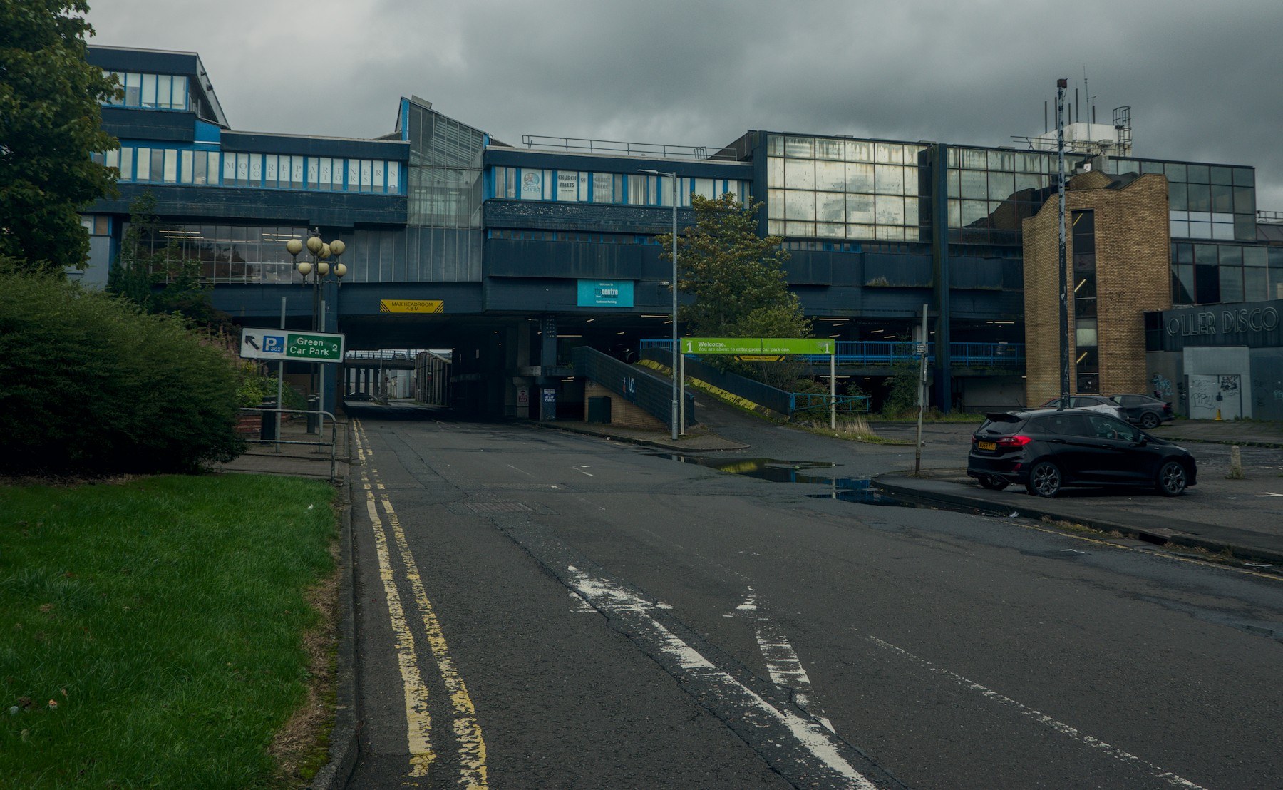Rain-darkened road approaching a blue and grey multi-level section of Cumbernauld Center, with a covered passage, ramp, parked black car and cloudy sky.