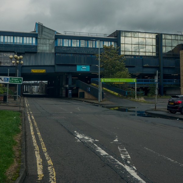 Rain-darkened road approaching a blue and grey multi-level section of Cumbernauld Center, with a covered passage, ramp, parked black car and cloudy sky.