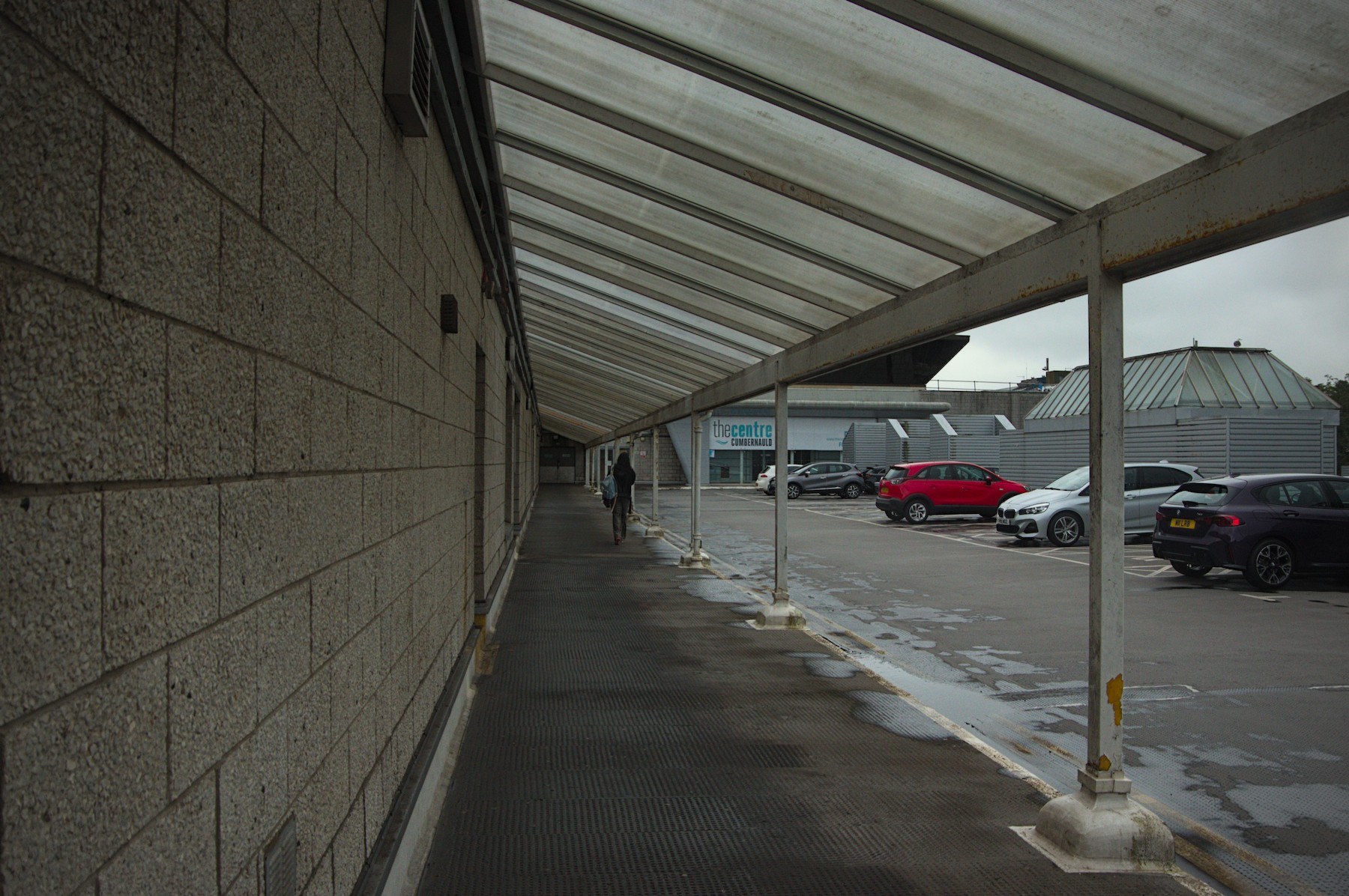 A pedestrian walks beneath a translucent covered walkway beside a concrete block wall, with wet paving and parked cars outside Cumbernauld Centre.