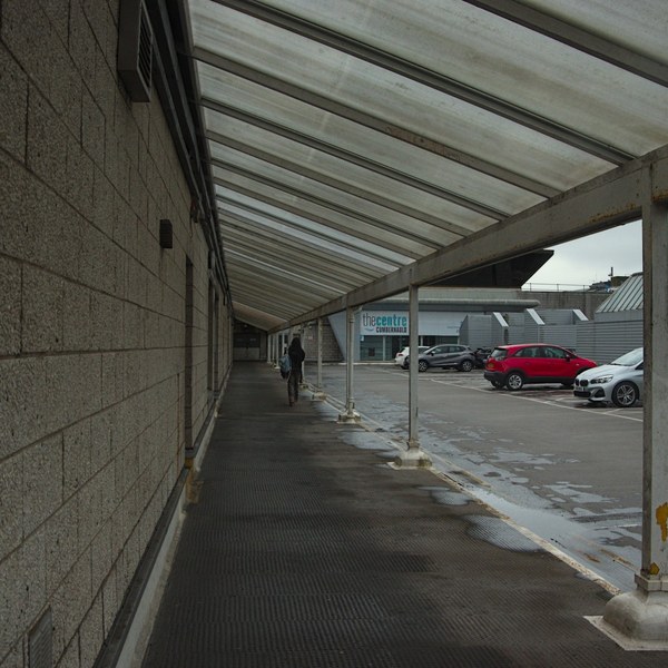 A pedestrian walks beneath a translucent covered walkway beside a concrete block wall, with wet paving and parked cars outside Cumbernauld Centre.