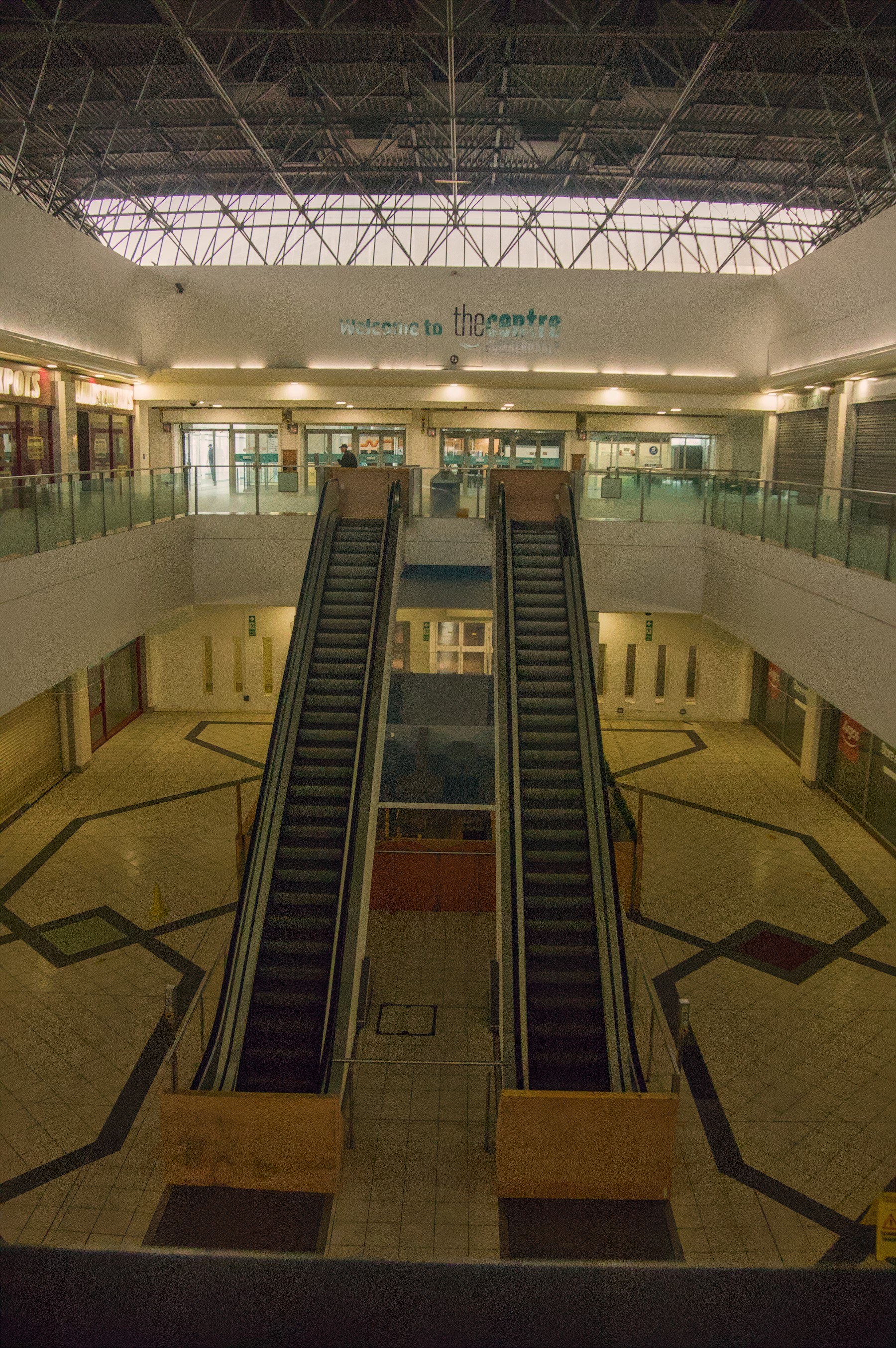 Two parallel escalators descend into an empty tiled shopping-centre atrium beneath exposed steel roof trusses; a “Welcome to the centre” sign is visible on the upper wall.