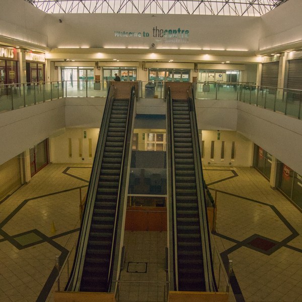 Two parallel escalators descend into an empty tiled shopping-centre atrium beneath exposed steel roof trusses; a “Welcome to the centre” sign is visible on the upper wall.