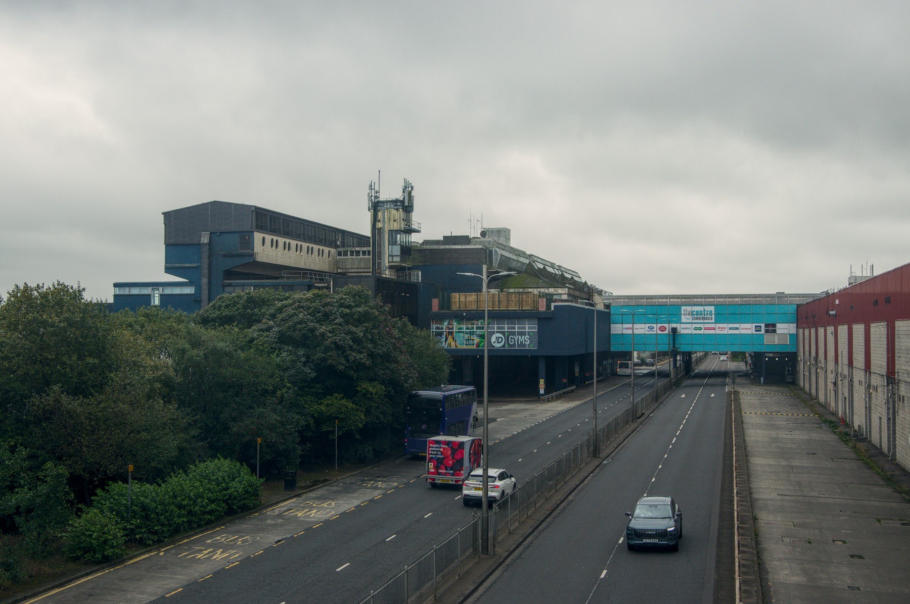 View down a divided access road at Cumbernauld Centre, with a bus and cars travelling toward commercial buildings, trees on the left and a signed bridge spanning the road ahead.