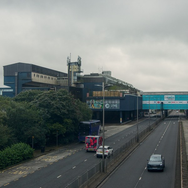 View down a divided access road at Cumbernauld Centre, with a bus and cars travelling toward commercial buildings, trees on the left and a signed bridge spanning the road ahead.