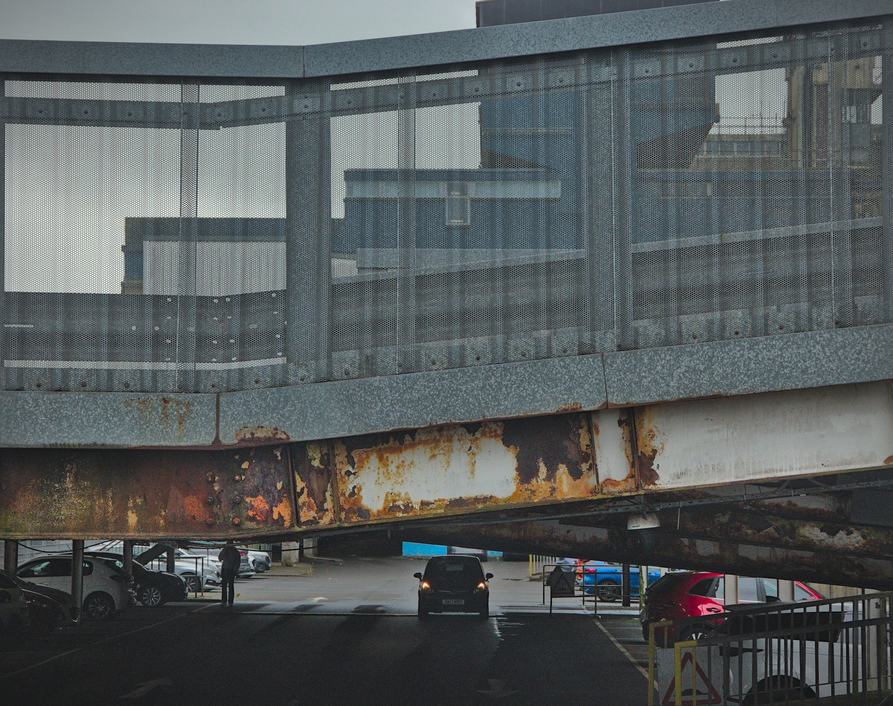 A van drives under a glazed pedestrian bridge with a heavily rusted steel beam; parked vehicles and a person are visible beyond.