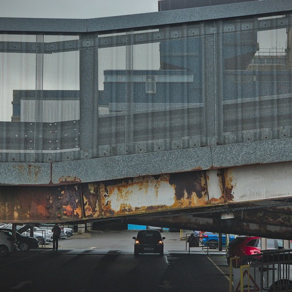 A van drives under a glazed pedestrian bridge with a heavily rusted steel beam; parked vehicles and a person are visible beyond.