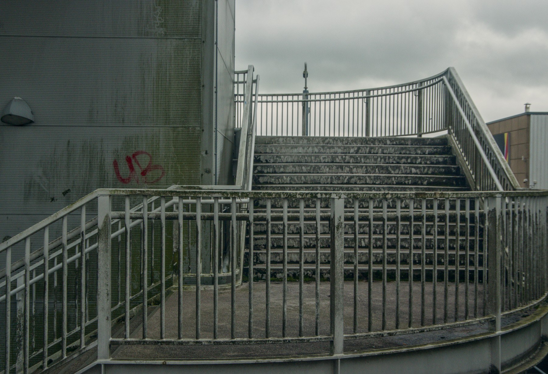 Exterior stairway with weathered metal railings, stained steps, a perforated wall with red graffiti, and grey clouds overhead.