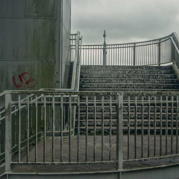 Exterior stairway with weathered metal railings, stained steps, a perforated wall with red graffiti, and grey clouds overhead.