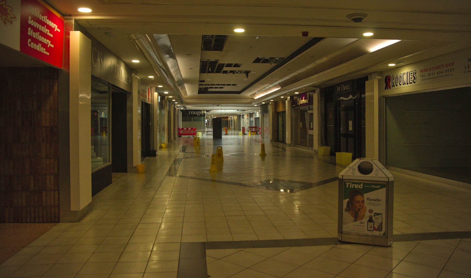 Empty shopping-centre arcade with closed shutters, yellow warning cones, wet tiled floor and a drinks advertising stand under ceiling lights.