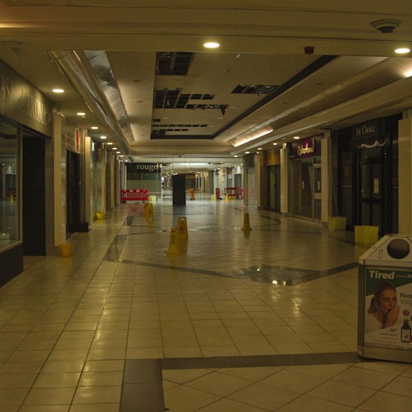 Empty shopping-centre arcade with closed shutters, yellow warning cones, wet tiled floor and a drinks advertising stand under ceiling lights.