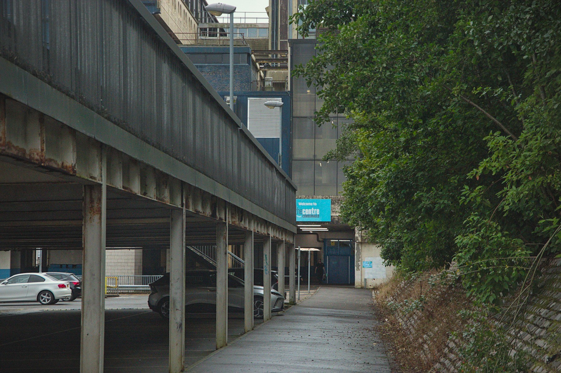 Wet pavement beside a covered car park at Cumbernauld Centre, with parked cars under steel supports, dense trees on the right, and a turquoise welcome sign ahead.