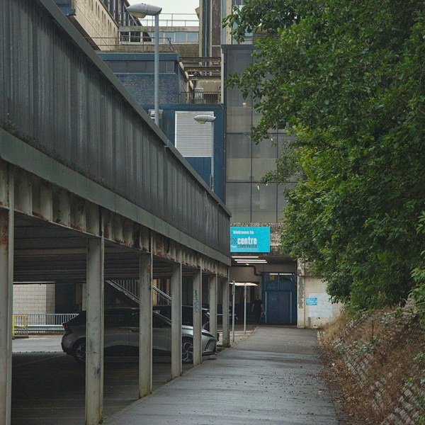 Wet pavement beside a covered car park at Cumbernauld Centre, with parked cars under steel supports, dense trees on the right, and a turquoise welcome sign ahead.