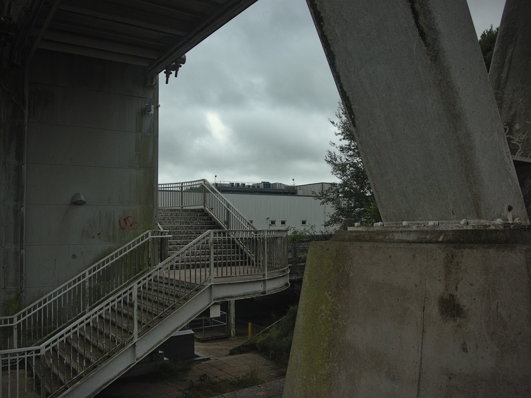 Metal pedestrian stairs and railings beneath a concrete canopy, framed by a large angled concrete support and grey sky.