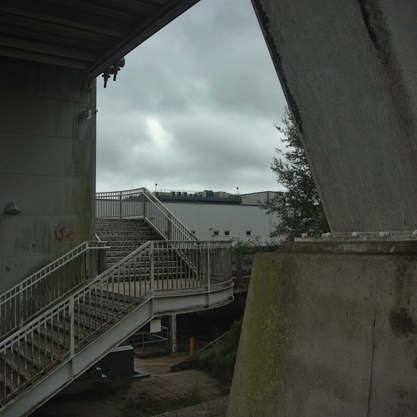 Metal pedestrian stairs and railings beneath a concrete canopy, framed by a large angled concrete support and grey sky.