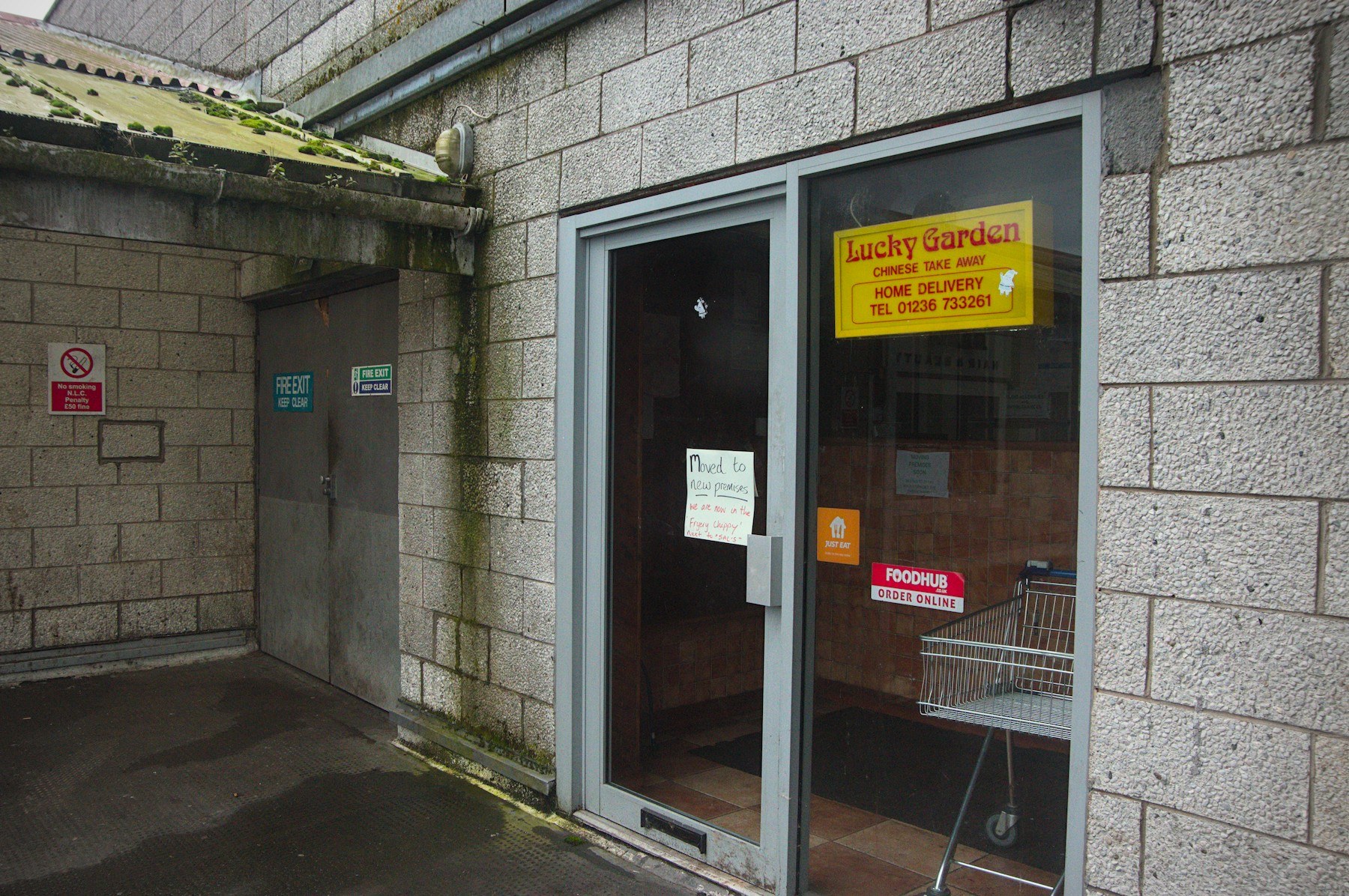 Glass entrance to Lucky Garden Chinese Take Away in a grey concrete passage, with delivery stickers, a handwritten notice and a shopping trolley inside.
