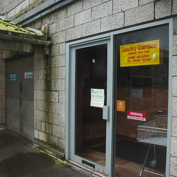 Glass entrance to Lucky Garden Chinese Take Away in a grey concrete passage, with delivery stickers, a handwritten notice and a shopping trolley inside.