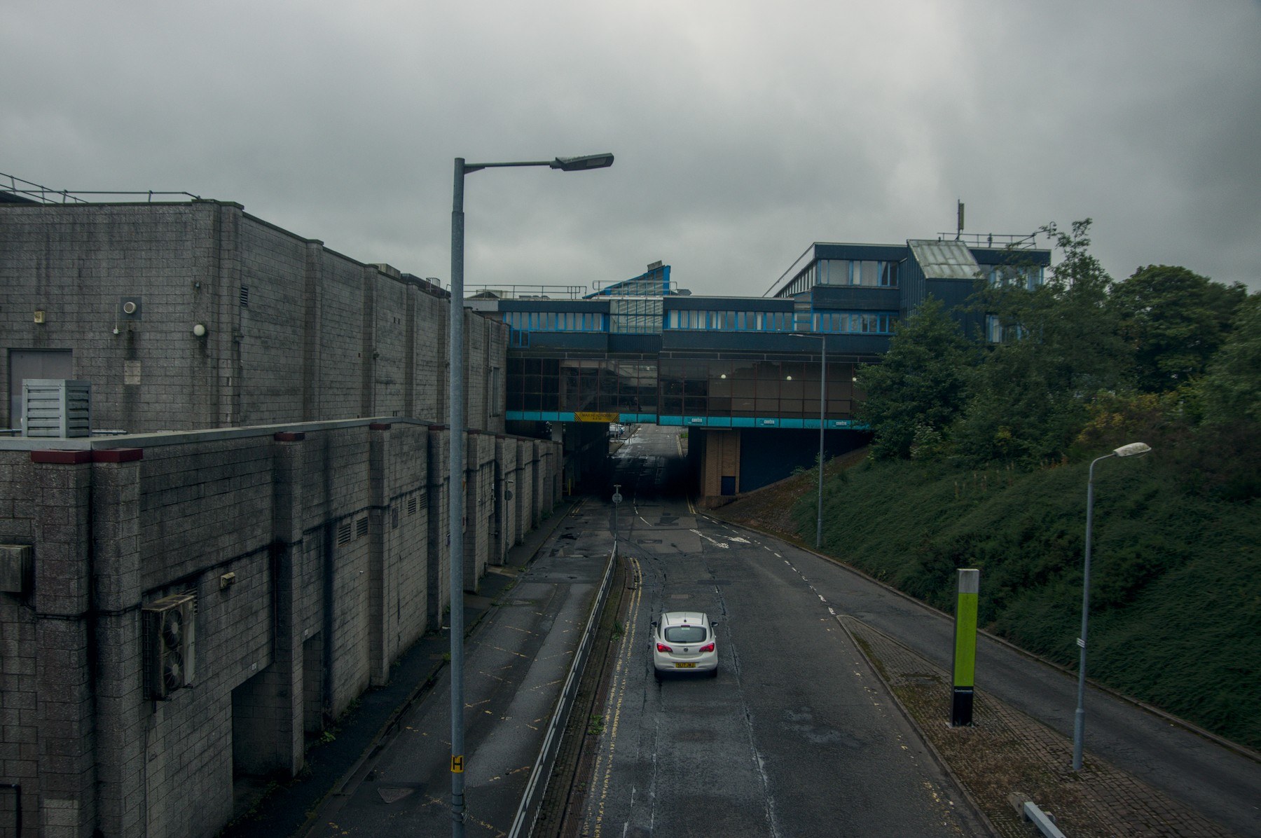 White car on a damp roadway between concrete walls and a grassy embankment, approaching a dark underpass below a blue-trimmed building.
