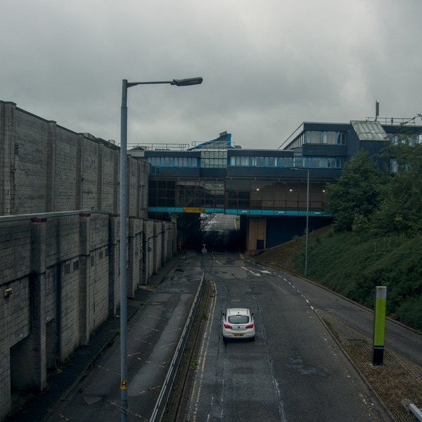 White car on a damp roadway between concrete walls and a grassy embankment, approaching a dark underpass below a blue-trimmed building.