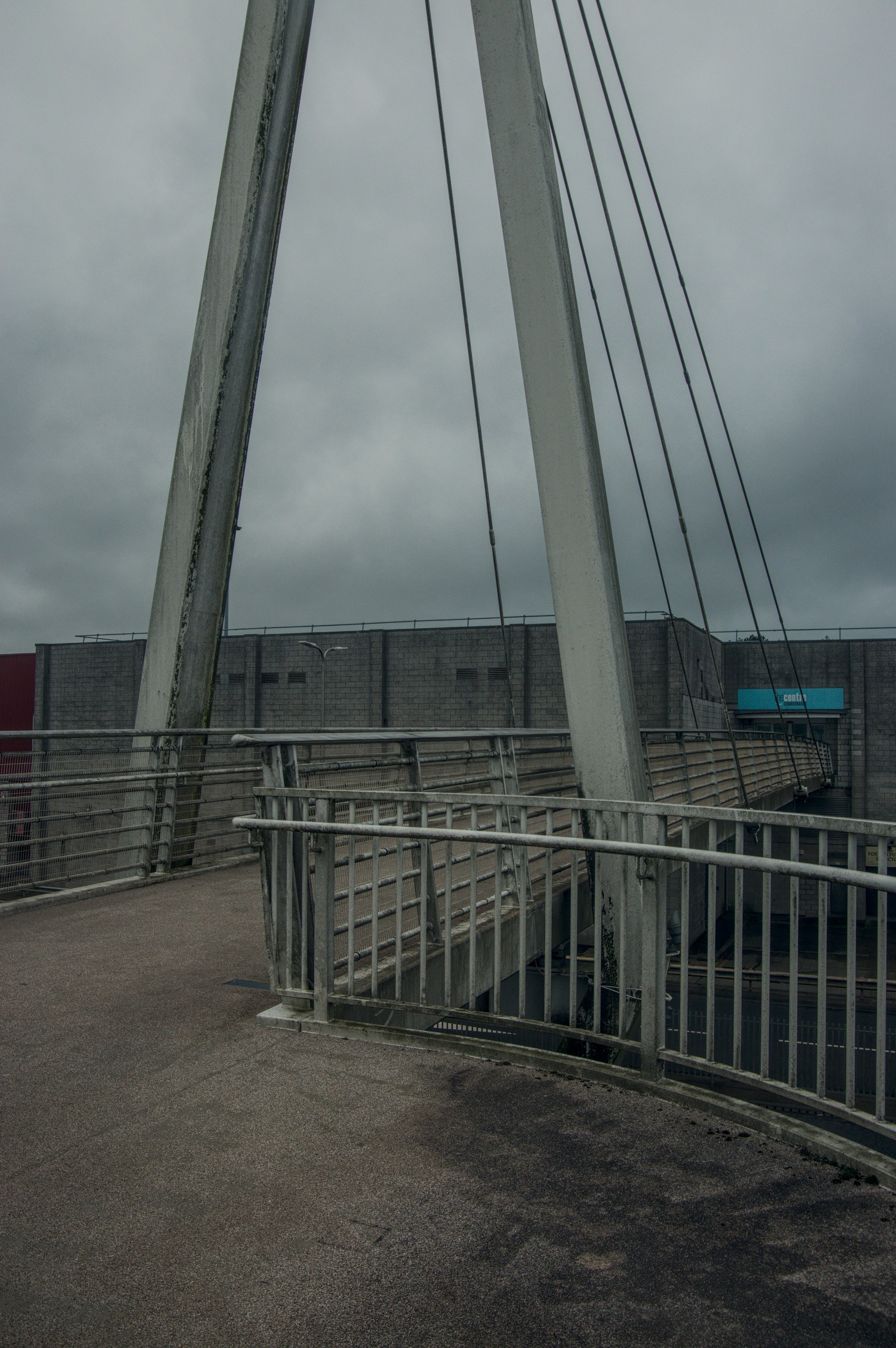 Curving pedestrian footbridge with metal railings, angled concrete supports and suspension cables beside a brick shopping-centre building under an overcast sky.