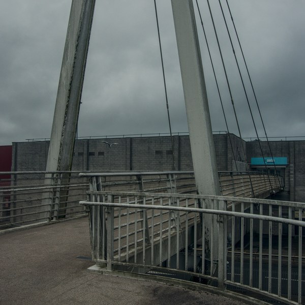Curving pedestrian footbridge with metal railings, angled concrete supports and suspension cables beside a brick shopping-centre building under an overcast sky.
