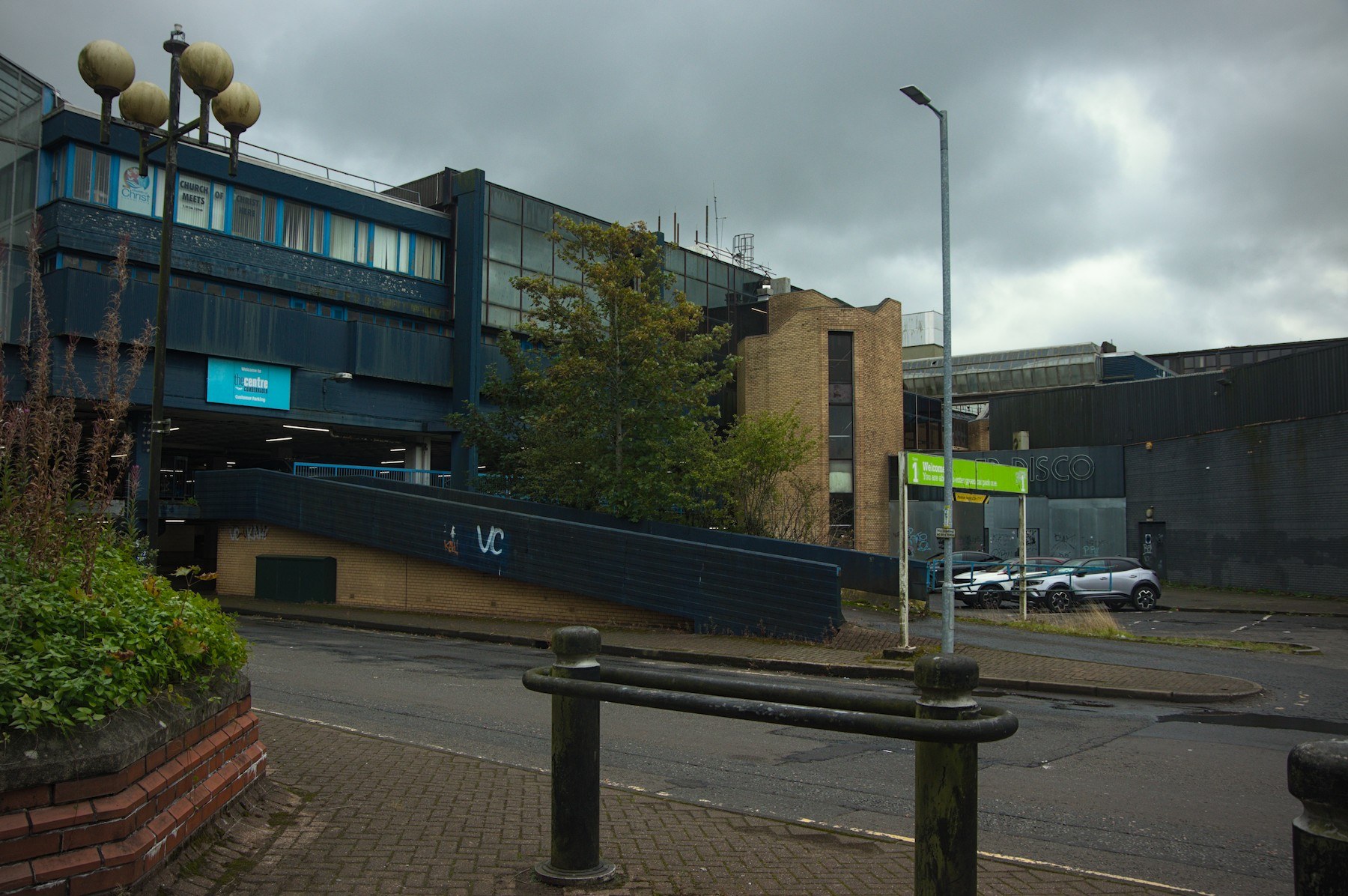Wet street outside Cumbernauld Center, with black bollards in the foreground, a ramp marked VC, parked cars and layered concrete and blue-panelled buildings under grey clouds.
