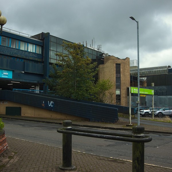 Wet street outside Cumbernauld Center, with black bollards in the foreground, a ramp marked VC, parked cars and layered concrete and blue-panelled buildings under grey clouds.