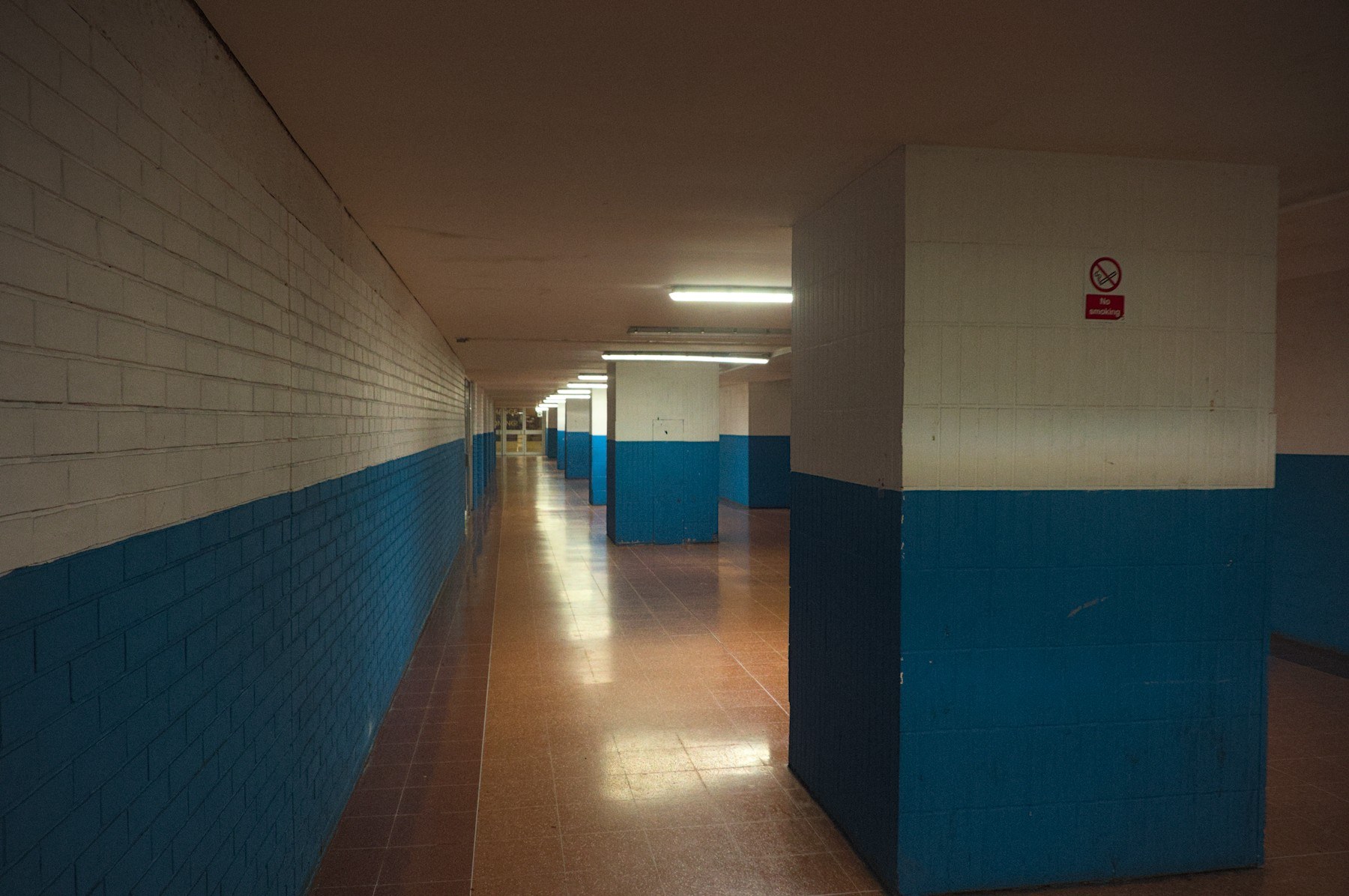 Empty tiled corridor in Cumbernauld Center with white upper walls, dark teal lower walls, fluorescent ceiling lights, and a no-smoking sign on a column.