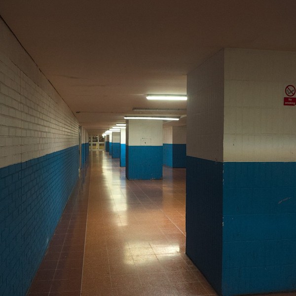 Empty tiled corridor in Cumbernauld Center with white upper walls, dark teal lower walls, fluorescent ceiling lights, and a no-smoking sign on a column.