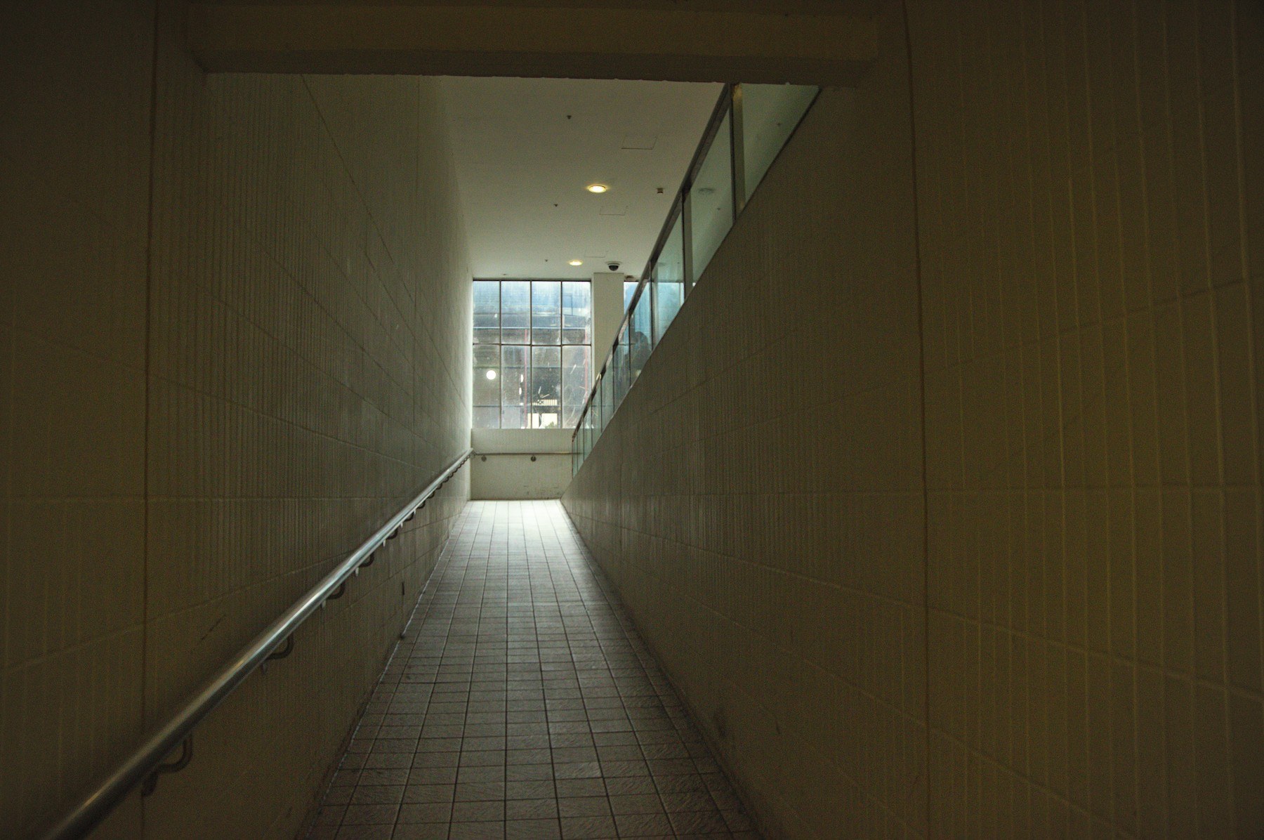 Tiled interior ramp with a metal handrail on the left, rising toward large windows and daylight at Cumbernauld Center.