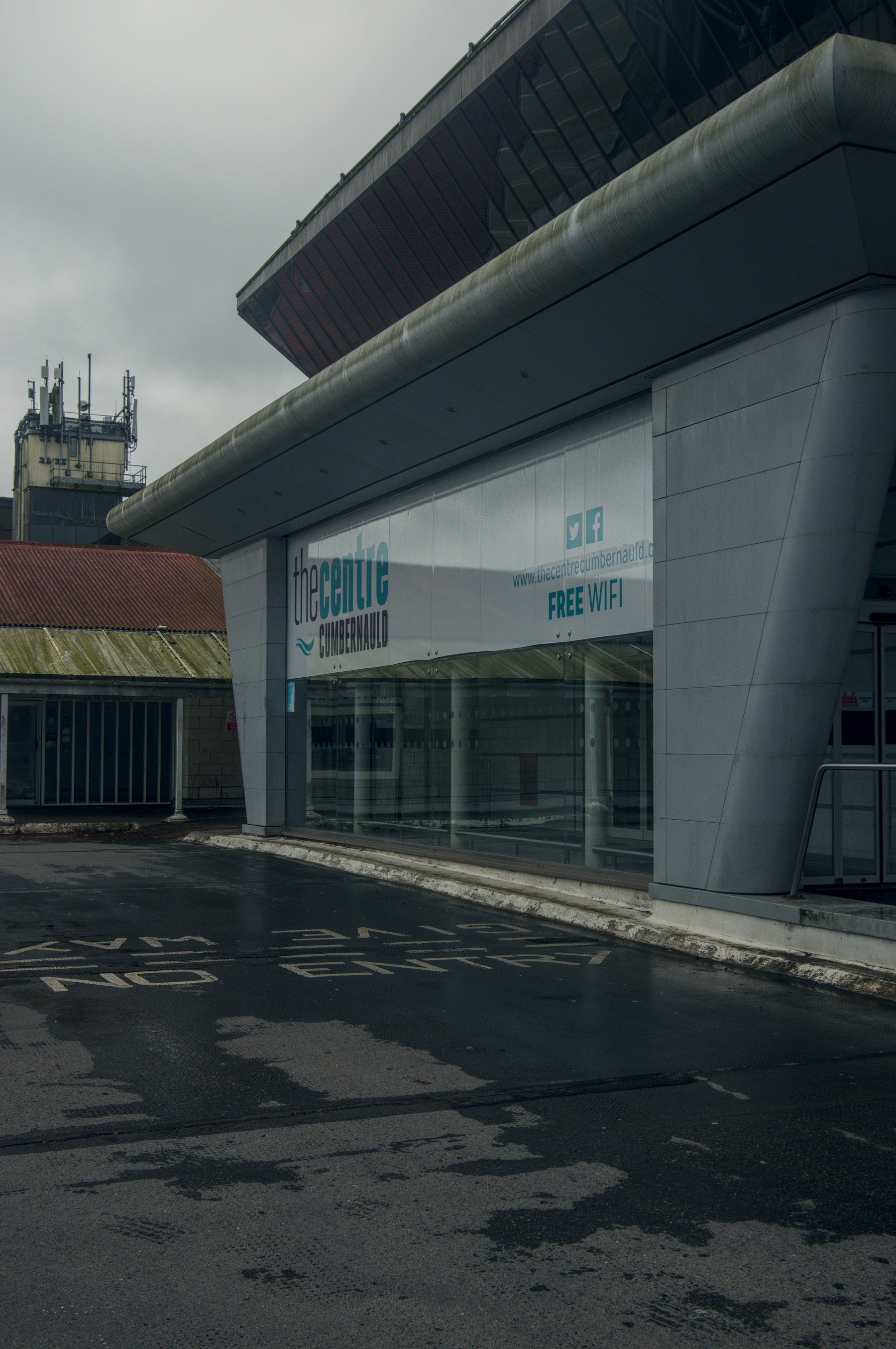 The Centre Cumbernauld entrance with a Free WiFi banner, sloping metal overhang, curved grey support column, and worn road markings on damp pavement.