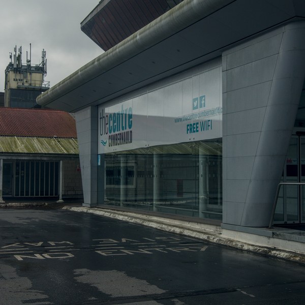 The Centre Cumbernauld entrance with a Free WiFi banner, sloping metal overhang, curved grey support column, and worn road markings on damp pavement.