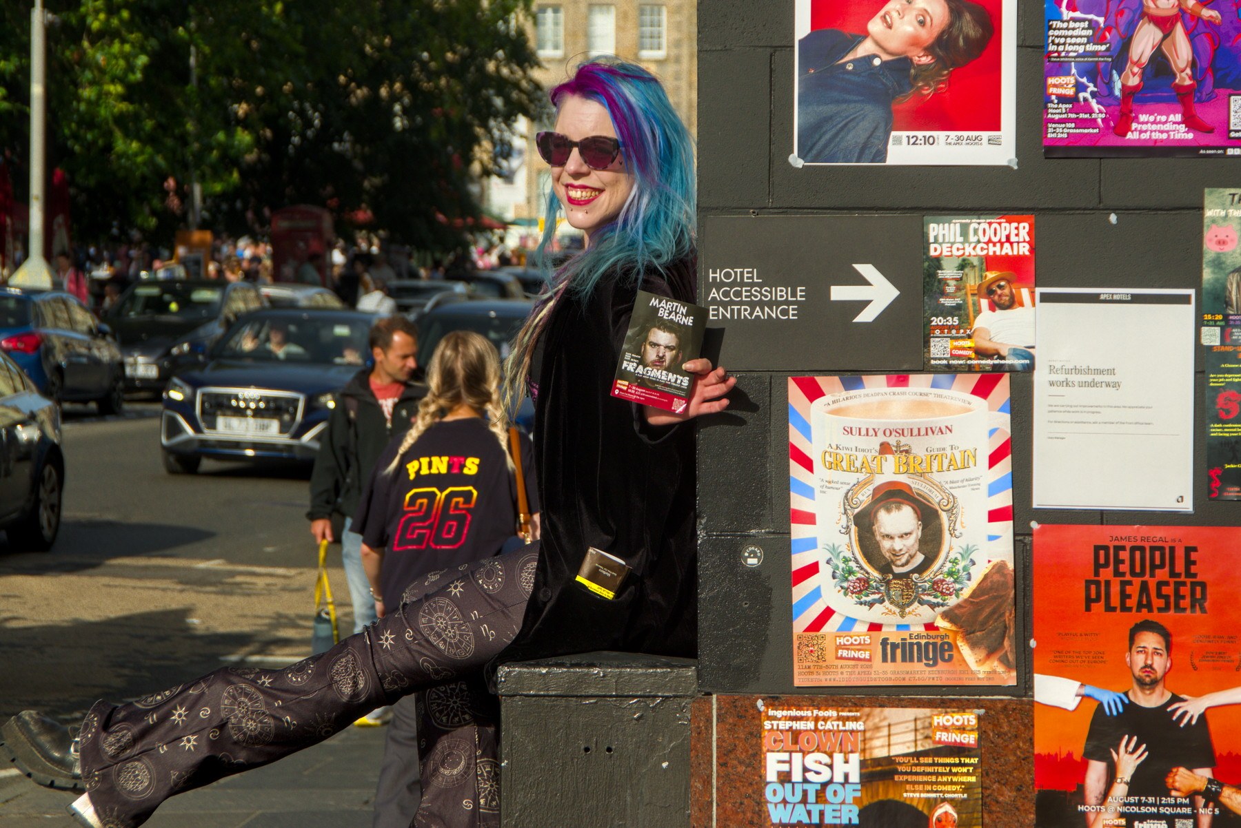 Blue- and purple-haired person in sunglasses smiles while holding a festival flyer beside a wall of Edinburgh Fringe posters and an accessible entrance sign.
