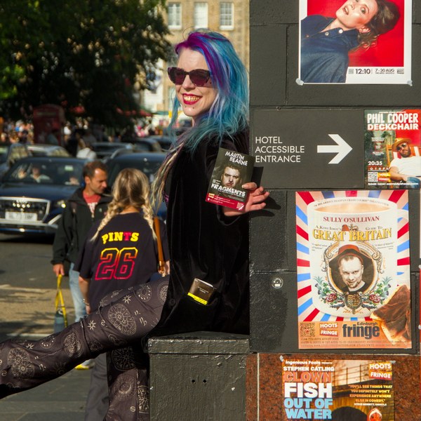 Blue- and purple-haired person in sunglasses smiles while holding a festival flyer beside a wall of Edinburgh Fringe posters and an accessible entrance sign.