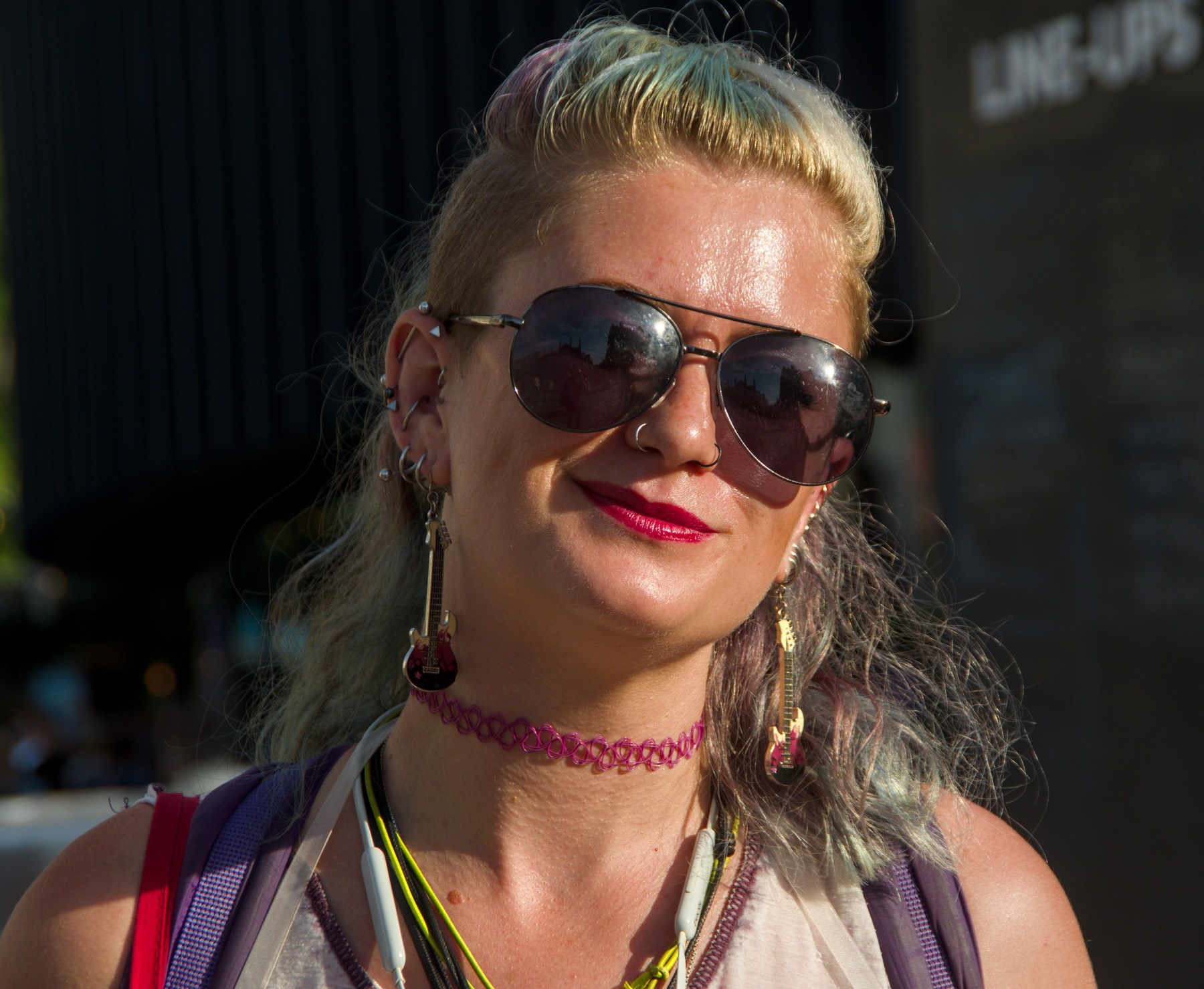 Close portrait of a smiling woman with aviator sunglasses, red lipstick, multiple earrings, a pink choker and backpack straps in Edinburgh city centre.