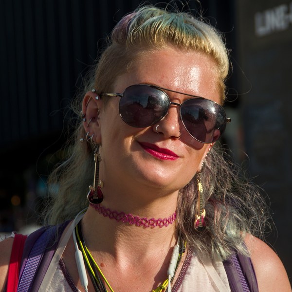 Close portrait of a smiling woman with aviator sunglasses, red lipstick, multiple earrings, a pink choker and backpack straps in Edinburgh city centre.