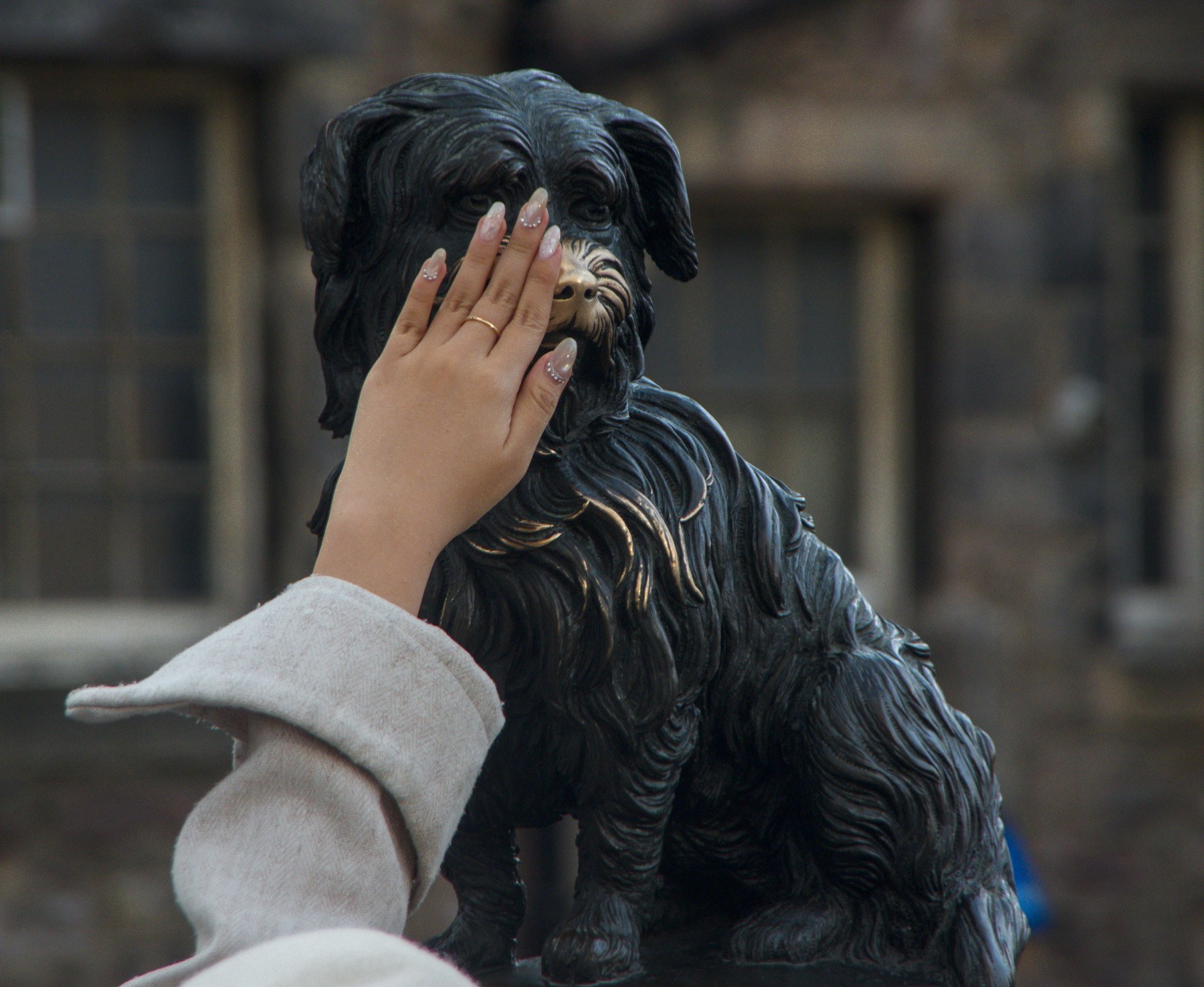 A hand with glittered nails rests across the face of a dark bronze dog sculpture, with a stone building blurred behind.
