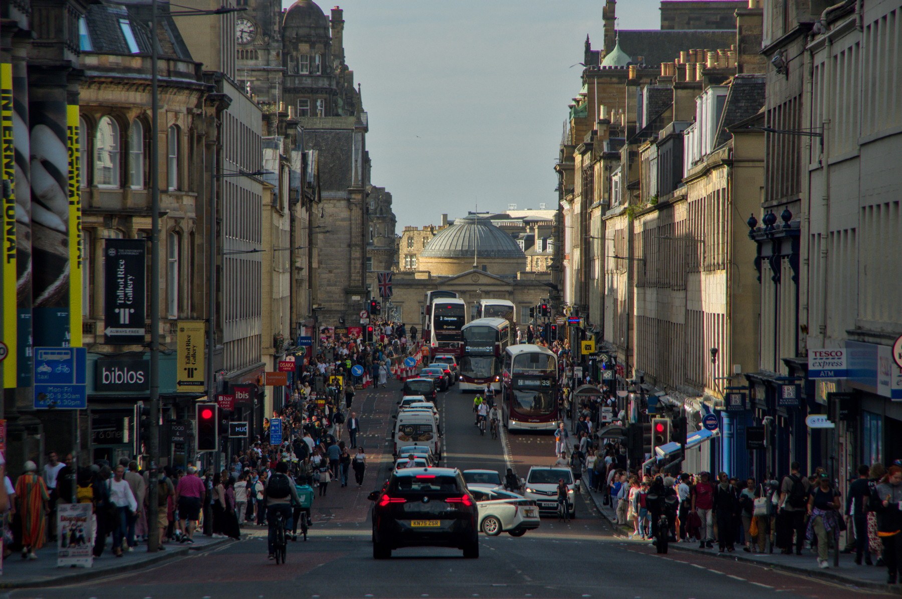 Elevated view down a busy Edinburgh city-centre street, with buses, cars, cyclists and dense pedestrian pavements leading toward a domed building.