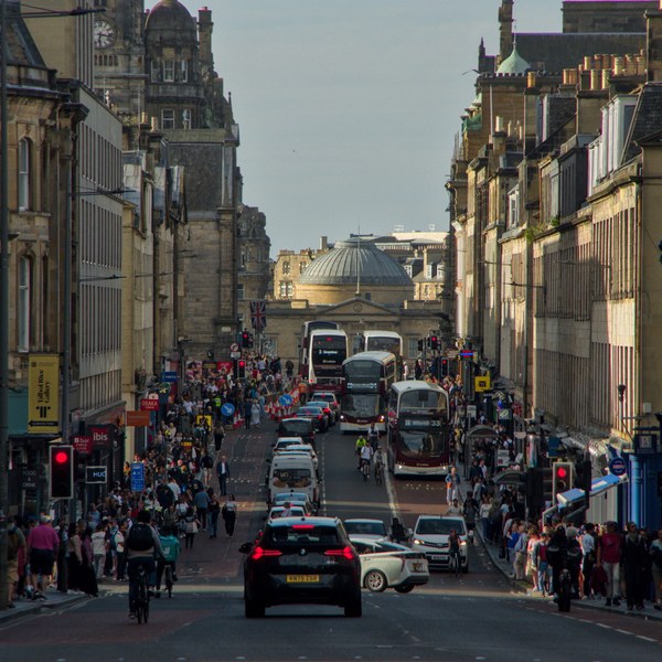 Elevated view down a busy Edinburgh city-centre street, with buses, cars, cyclists and dense pedestrian pavements leading toward a domed building.