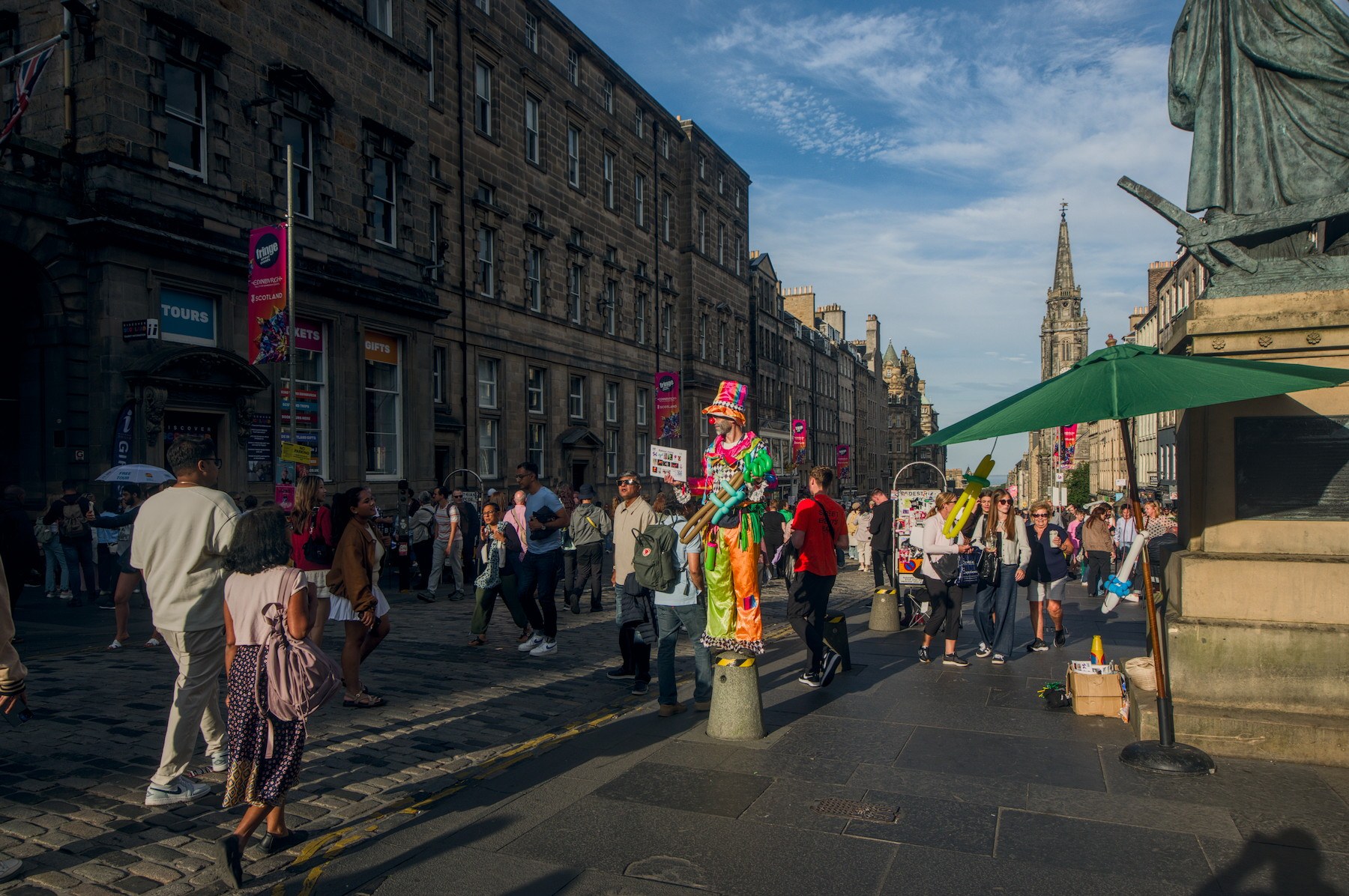 Colourfully costumed street performer stands among pedestrians on a historic Edinburgh street, with a green parasol and church spire behind.