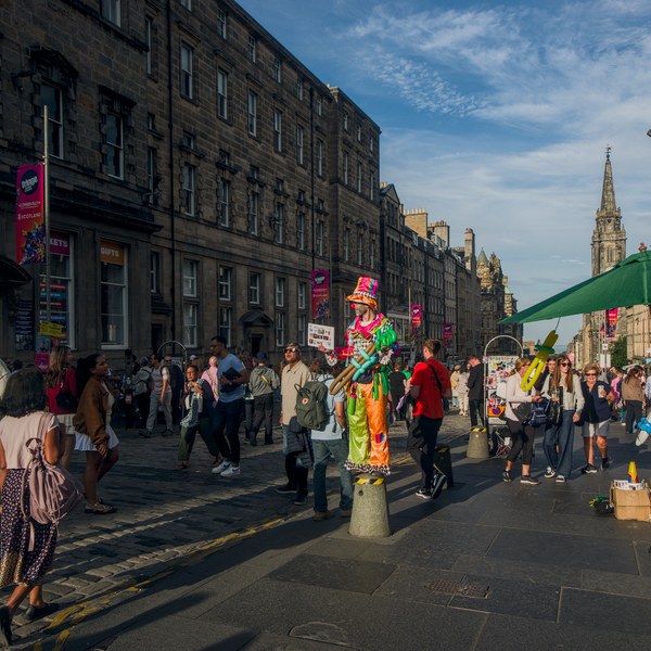 Colourfully costumed street performer stands among pedestrians on a historic Edinburgh street, with a green parasol and church spire behind.