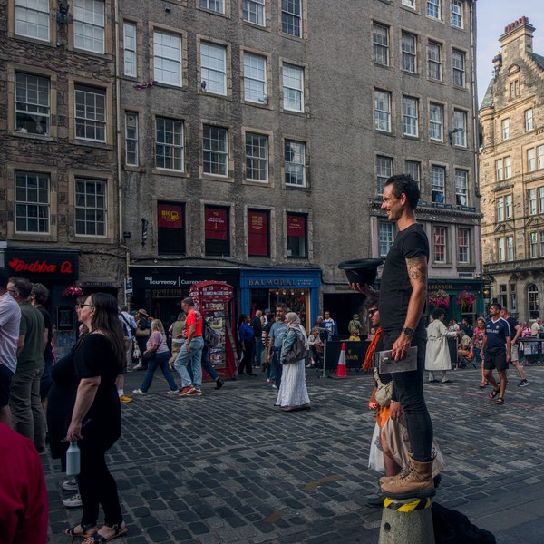 A man in black stands on a short bollard holding a black hat above a crowd on a cobbled Edinburgh street, with tall stone buildings behind him.