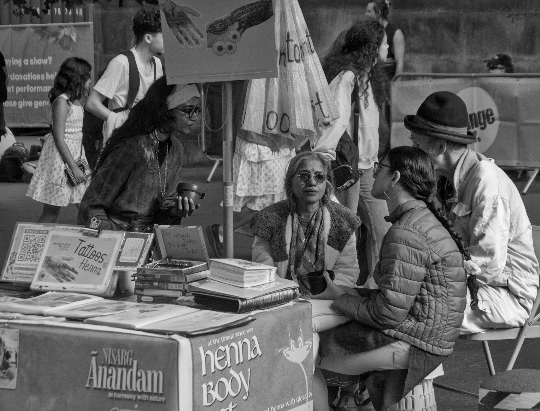 A henna artist seated behind a display of design books and a “Tattoos Henna” sign speaks with two visitors at a busy Edinburgh festival stall.