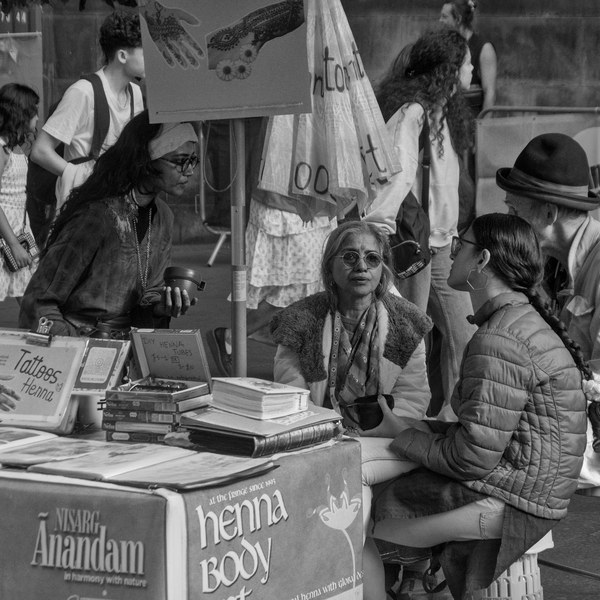 A henna artist seated behind a display of design books and a “Tattoos Henna” sign speaks with two visitors at a busy Edinburgh festival stall.
