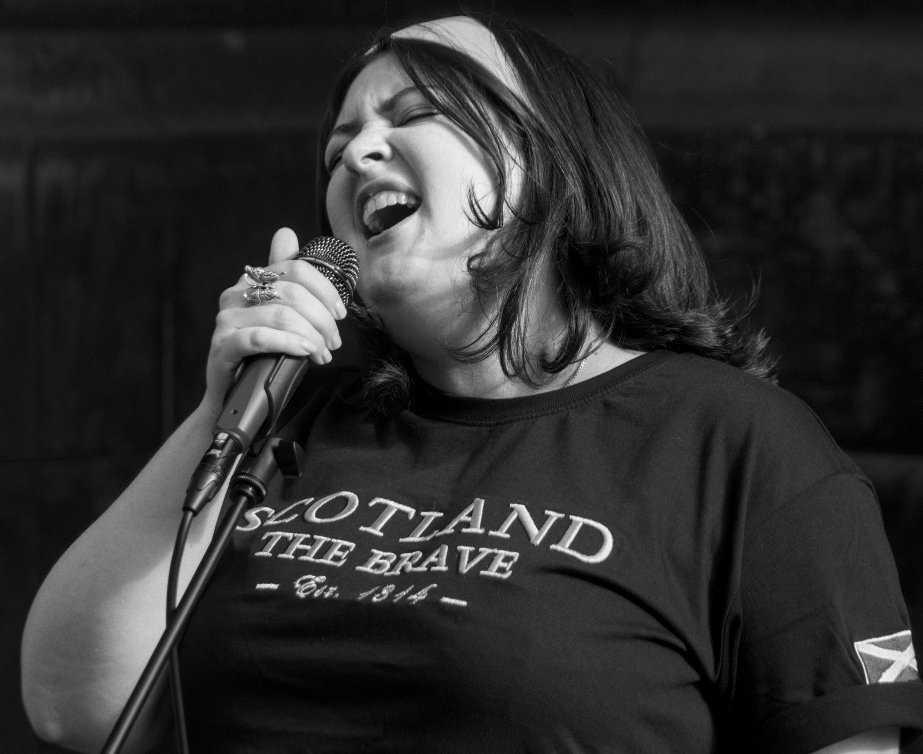 Black-and-white close-up of a woman singing into a wired microphone, wearing a T-shirt printed “Scotland the Brave.”